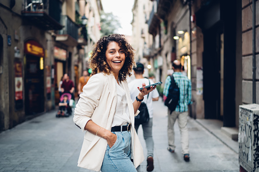 Side view of young stylish ethnic lady in casual clothes standing with smartphone on narrow pedestrian street of old city and looking at camera