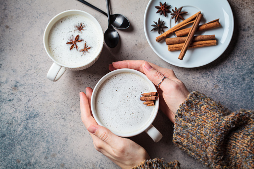 Homemade chai latte with cinnamon and star anise in white cup in hands, dark background, top view.