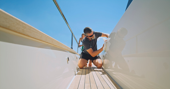 Yacht cabin crew cleaning luxury yacht while kneeling against clear sky.