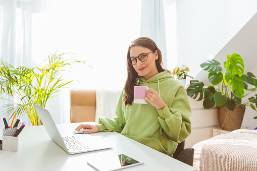 Beautiful woman wearing green hoodie, sitting at the table in her bedroom and using laptop, smiling at camera. Home office concept.