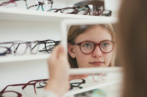 Girl holding a mirror and looking at different new eyeglasses to buy. Trying new models and testing them in the mirror.