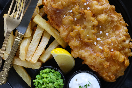 Stock photo showing elevated view of a black plate filled with a portion of battered cod fish and chips, with lemon slices and garnished with parsley, besides ramekins of mushy peas and tartare sauce, salt and pepper mills and knife and fork.