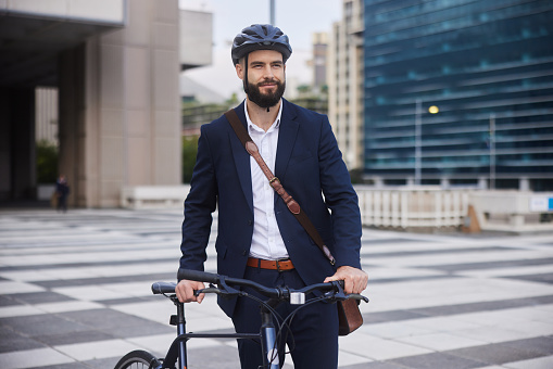 Smiling young businessman with a bag wearing a cycling helmet and walking with his bike after leaving work