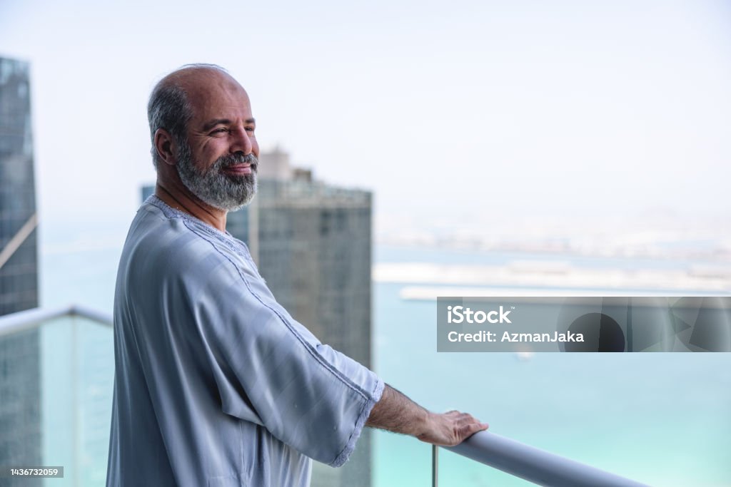 Middle Eastern Older Male Enjoying View On His Balcony Middle eastern older male is wearing traditional clothes and enjoying beautiful view on his balcony. Large buildings in the background. Islam Stock Photo