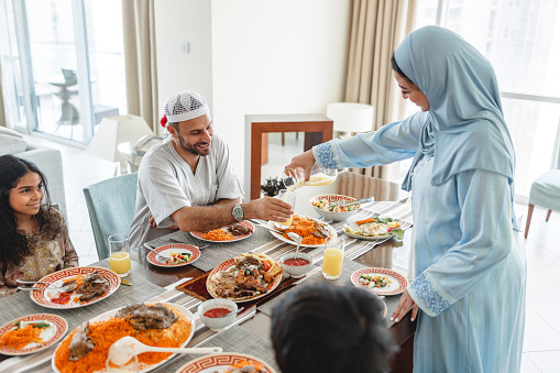Middle eastern wife is pouring orange juice to her husband. They are having family lunch with their children. They are all wearing traditional clothes.