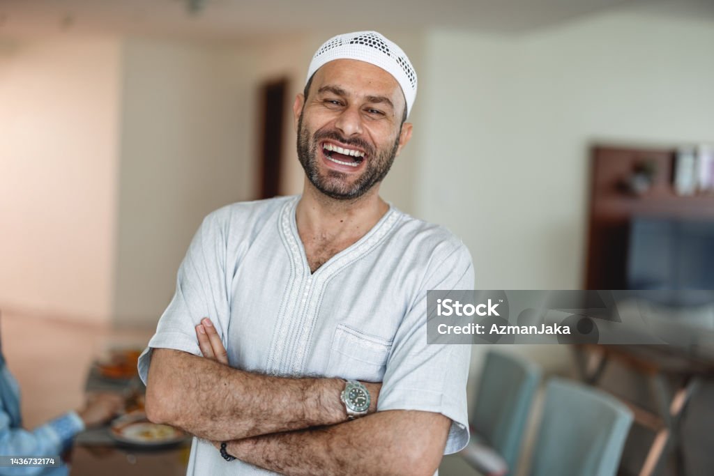 Close Up Portrait Of Middle Eastern Middle Age Male Laughing Close up portrait of middle eastern age male is laughing while standing cross-armed in dining room. He is wearing grey traditional clothes and white muslim cap. Men Stock Photo