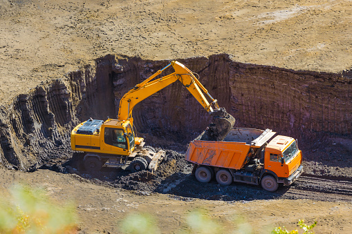 Yellow excavator loading an orange dump heavy truck with soil during quarrying. Construction vehicles working in soil quarry. Excavator digs the ground. Top view