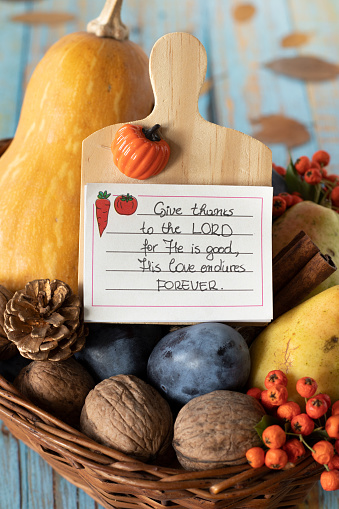 Thanksgiving day card with handwritten text for giving thanks to the LORD in a wicker basket with autumn fruit on a wooden table. Vertical shot, a close-up. Christian gratitude and praise concept.