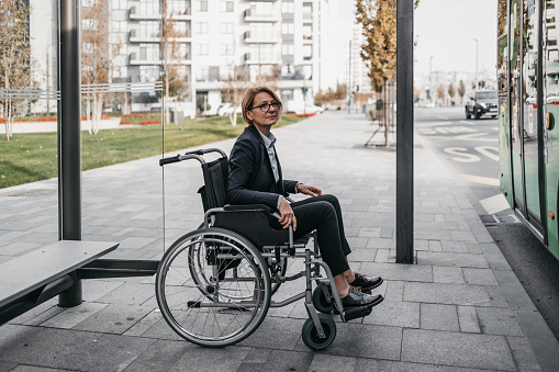 Disabled senior business woman in a wheelchair is waiting for city transportation on street bus stop.