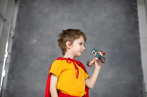 Dreamy boy in a red cape plays with an airplane toy on a gray artistic background. Little super hero.