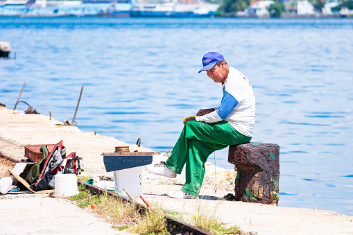 Havana, Cuba - September 15, 2022: An older man sitting on a rock cross-legged beside the sea. The man is wearing a baseball cap, and some fishing gear is on the scene.