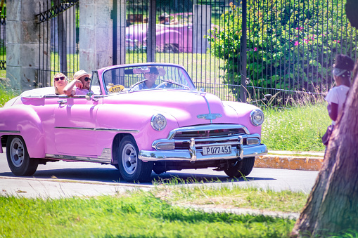Havana, Cuba - September 15, 2022: Three people traveling in a pink convertible car on a sunny day. There is grass on the sidewalk of the road, and a metal fence is in the background.