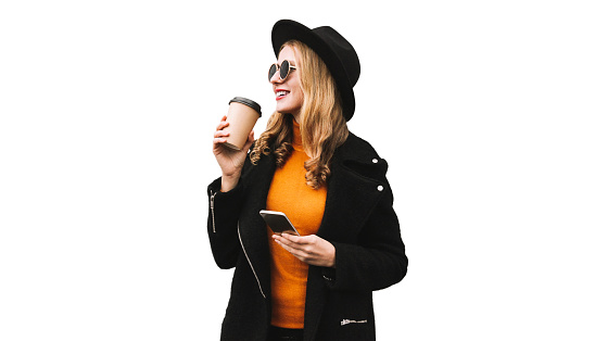 Stylish portrait of beautiful young woman drinking coffee with smartphone looking away wearing black coat and round hat isolated on white background