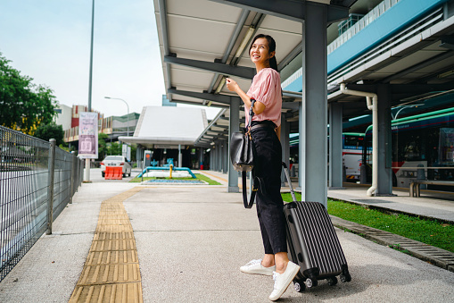 Asian woman walking on sidewalk beside near bus station and pulling wheeled luggage
