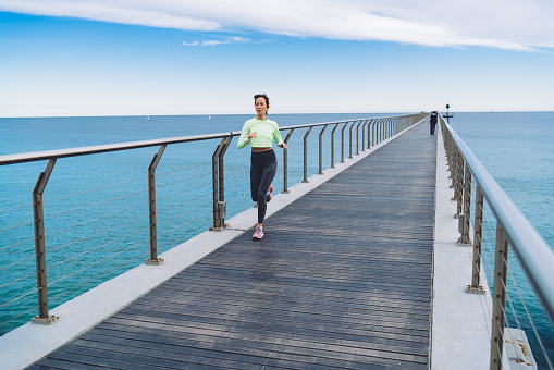 Full body of slim adult female in sport leggings and top jogging on wooden pier against waterfront and blue sky