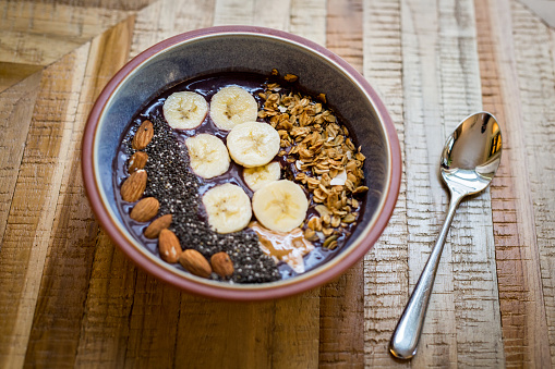 Acai Bowl made with frozen açai, coconut milk, yogurt, slices of banana, chia seeds, granola, almonds in a bowl on the table with a spoon