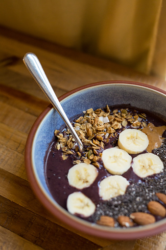 Acai bowl made with Acai, coconut milk, sliced bananas, granola, almonds, chia seeds, and peanut butter in a bowl on a table with a spoon