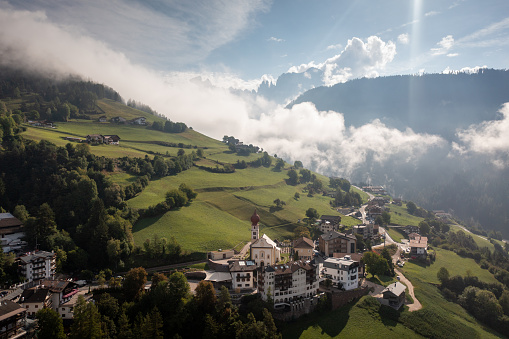 A lovely village with a majestic church in the stunning surroundings of the Dolomites mountains at sunrise. The low hanging clouds deliver soft texture and create an idyllic scene.