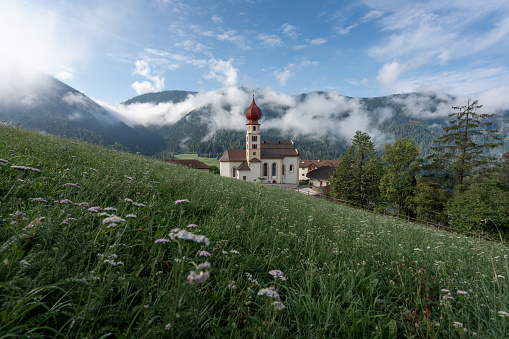 A lovely village with a majestic church in the stunning surroundings of the Dolomites mountains at sunrise. The low hanging clouds deliver soft texture and create an idyllic scene.