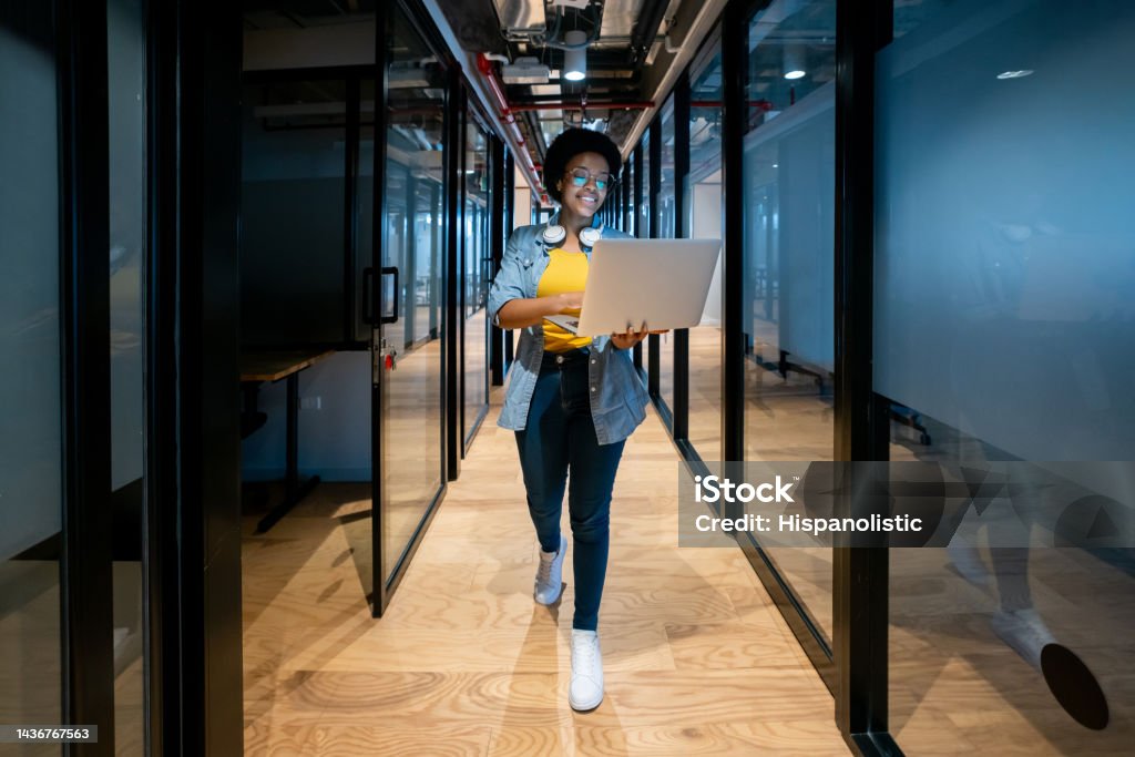 Happy IT technician working at the office using her laptop Happy African American IT technician working late at the office using her laptop and smiling Technology Stock Photo