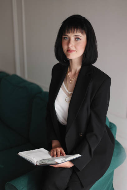 istock Beautiful business woman writing something in the note pad while sitting on the sofa at office, charming brunette female in black suit looking in camera, girl writes diary or work indoors 1436770624