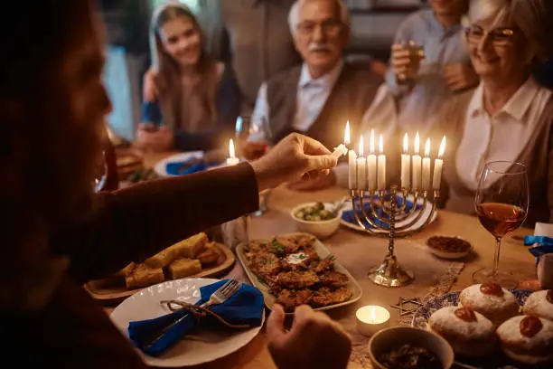 Close up of extended Jewish family celebrating Hanukkah while gathering at dining table. Focus is on father lighting the menorah.