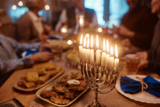 Close up of menorah on dining table with extended Jewish family in the background. Lit candles in menorah during family meal at dining table on Hanukkah. hanukkah candles stock pictures, royalty-free photos & images