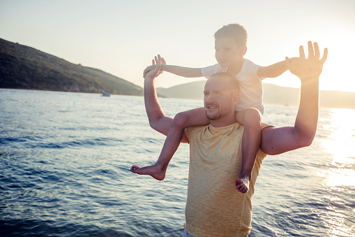 Cheerful father piggybacking son at sea shore. Happy man is playing with boy at beach on sunny day. They are enjoying summer vacation.