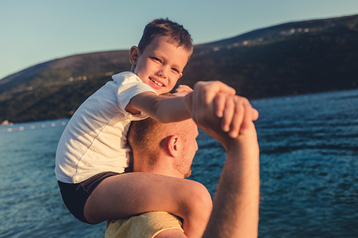 Portrait of happy caucasian father and playful son having fun in the sun against blue sky outside at sea. Happy father piggybacking his son at beach during sunny day.