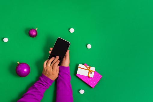 Hands in a purple sweater touches the smartphone screen with fingers, online payment for Christmas shopping, green background, flat lay. Bright concept of online shopping. Only hands in the frame