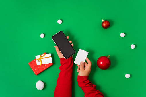 Female hands hold a smartphone and a bank card on a green background, Christmas decor and gifts. Flat lay, minimal Concept of online payment online shopping