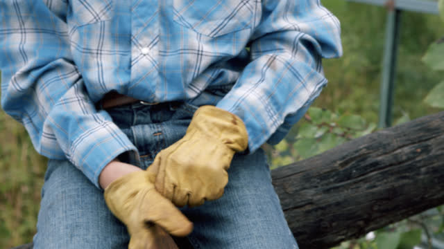 Young Caucasian American Rancher Child Leaning Against a Split Rail Fence Putting on His Leather Protective Work Gloves for a Hard Day of Work on a Small Town Family-Owned Ranch in Colorado, USA