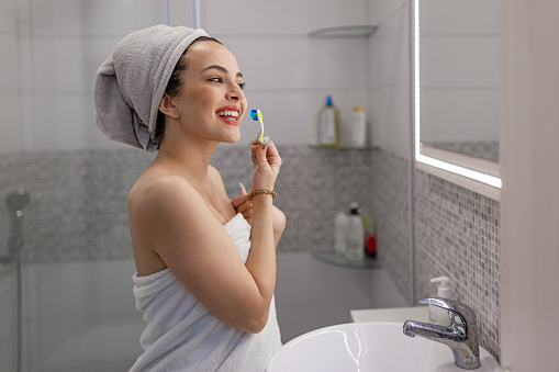 Beautiful smiling young Caucasian woman brushing her teeth in the morning in bathroom while looking herself in the mirror