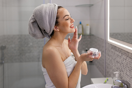 Adorable healthy young Caucasian woman taking care of her skin, applying a moisturizer in the bathroom