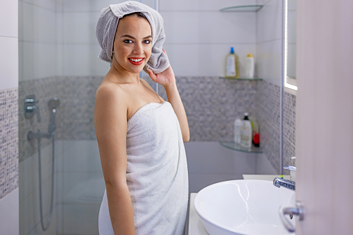 Beautiful cheerful young Caucasian woman standing wrapped in a towels and smiling for the camera in bathroom