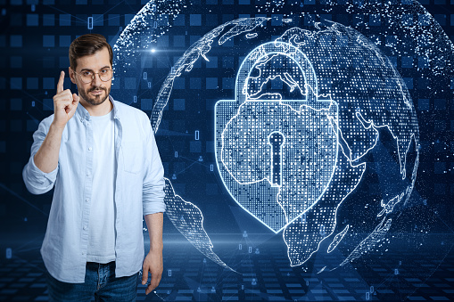 Attractive young european guy pointing up at abstract glowing padlock and globe hologram on blurry blue background. Global web safety and protection concept