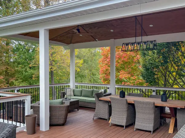 A luxury elevated deck with wicker patio furniture, wood dining table with chairs, ceiling fan and modern chandelier.  Pictured in autumn with fall colors.