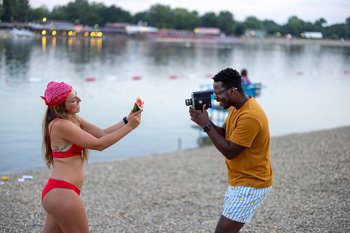 An African man is using an old camera to take a photo of his female friend eating watermelon while standing near the river or sea.