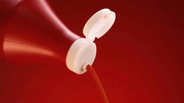 Photo of Ketchup, tomato sauce isolated on red background. Stock footage. Close up of ketchup pouring out of plastic bottle with white lid, concept of food preparation.