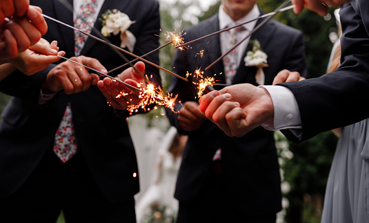 A crowd of young happy people in suits with sparklers in their hands during celebration. Sparkler on a wedding - bride, groom and guests holding lights in hand. Sparkling lights of bengal fires