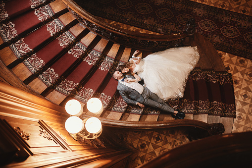 wedding day. beautiful bride in white long dress and young groom wearing in black suit are hugging and lying on the stairs inside big historic europe building.
