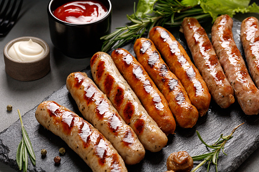 Stock photo showing close-up, elevated view of a portable kettle charcoal barbecue in a garden setting, stood on a wooden patio table featuring a large expanse of white, interconnecting, white plastic decking tiles in the background. Pictured cooking on the barbecue are some sausages and kebabs on wooden skewers made from chicken pieces and chorizo slices.