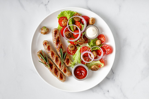Grilled barbecue sausages and vegetable salad with lettuce and tomatoes on white plate and marble background. Top view, copy space. BBQ