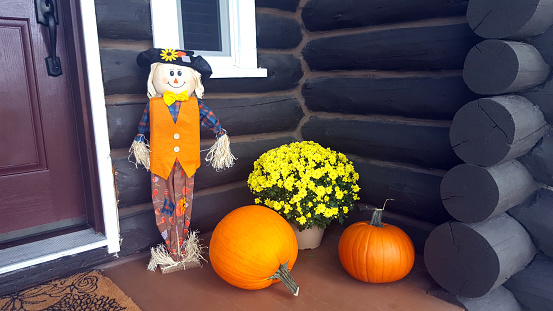 Vernon, BC, Canada- September 28,2022:  Front porch of old log cabin with Autumn arrangement of pumpkins,chrysanthemums, and straw man.  Partial door and window. Log structure.