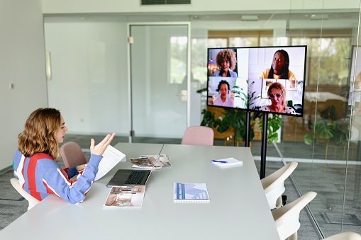 Business woman sitting in a meeting room with virtual attendees on the screen. Multiracial group of people on a video conference meeting in hybrid office space.