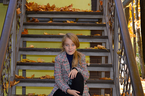 beautiful girl in a plaid coat on the stairs strewn with autumn leaves while walking in the park. the child enjoys the good weather, the beauty of autumn and a good mood