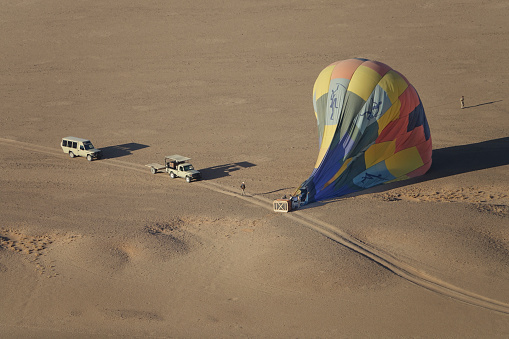 In September 2019, a recently landed hot air balloon and basket, both lying sideways, are attended to by personnel in the Namib Desert.  Balloons such as this one take passengers on scenic tours over a wilderness reserve in the desert,  which extends along the coast of Namibia.