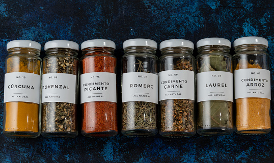 row of several glass jars, lying on a blue table. the jars are elongated. they have different seasonings, such as spicy, bay leaf, turmeric, seasoning for meat and for rice, provenzal.