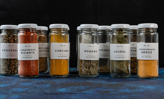 several glass jars with different condiments. the jars are elongated and have a white lid, they are on a blue table.