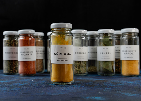 several glass jars with different condiments. the jars are elongated and have a white lid, they are on a blue table. close up of turmeric jar.